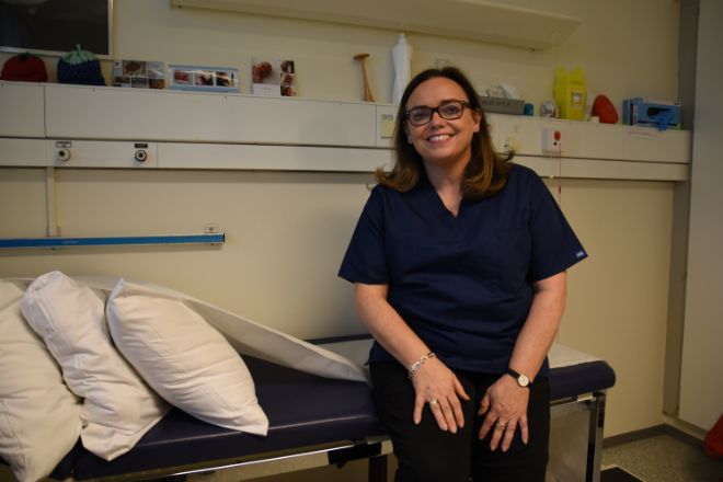 Fastlege og forsker Heidi Lidal Fidjeland i blå helseklær, sitter på en pasientbenk på legekontor.